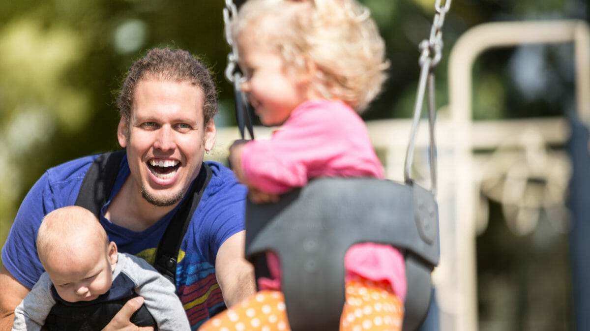A man holding a baby on his chest while pushing a young girl in a bucket swing.