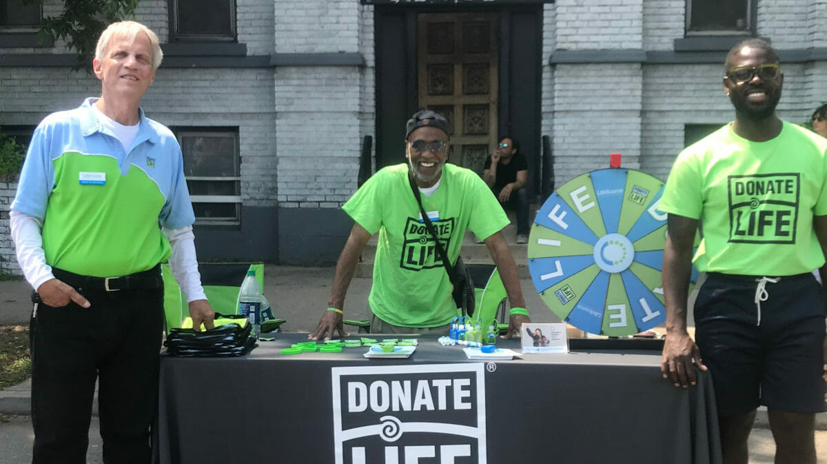 Three men standing at a Donate Life table with Donate Life swag sitting on the table.