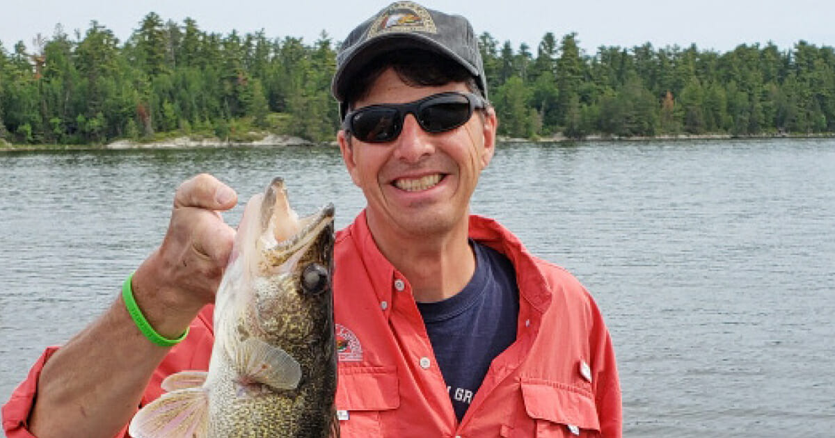 A man wearing a grey hat and sunglasses, smiling while holding a large fish by the mouth