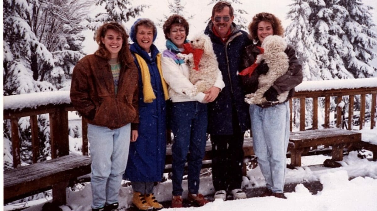 A family of 5 standing outside in the snow wearing winter jackets and smiling. Two white dogs are being held by two of the women.