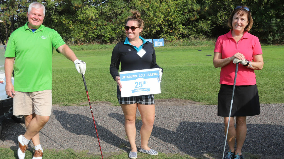 3 individuos de pie con palos dorados en un campo de golf. La mujer del centro sostiene un cartel de LifeSource Gold Classic 25th Anniversary.