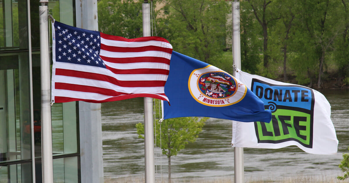 Bandera de Estados Unidos, bandera del estado Minnesota y bandera de Dona Vida en mástiles ondeando al viento.