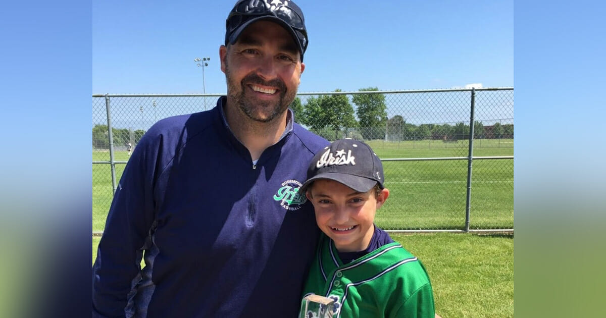 Un hombre con gorra de béisbol rodea con el brazo a un niño con uniforme de béisbol verde.