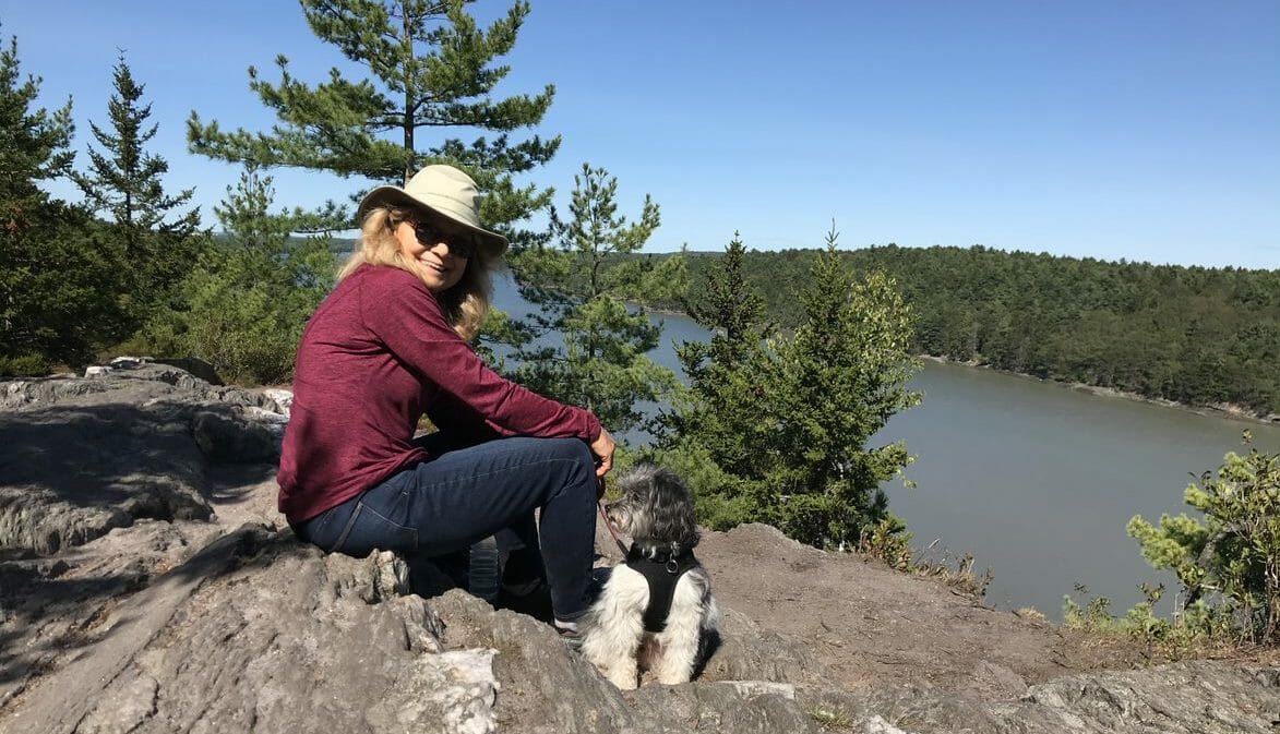 Joanie con camisa roja y sombrero marrón sentada en una roca al aire libre con su perro mirando un lago