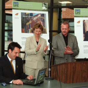 El gobernador Tim Pawlenty sentado en una mesa y utilizando un ordenador portátil. Dos personas junto a Tim aplauden.