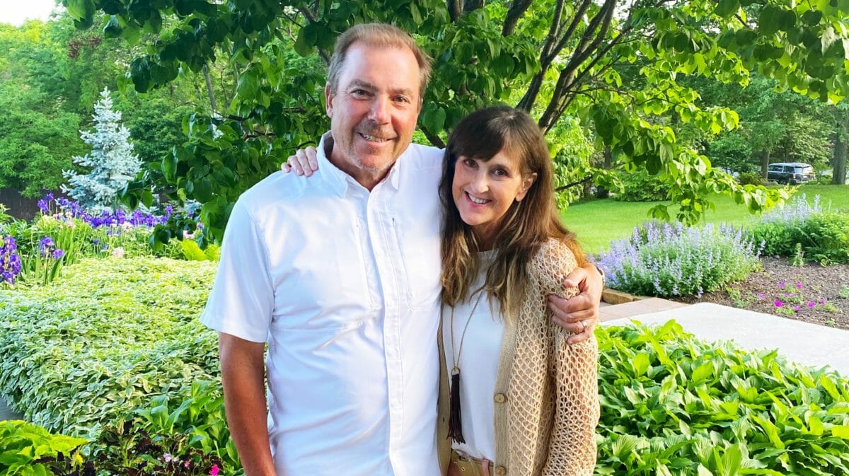 Jennifer CM wearing a brown sweater with her arm around her husband. Both are smiling