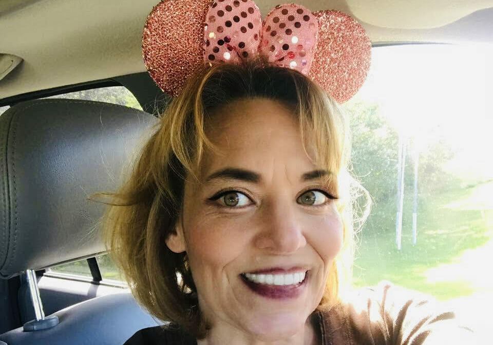 Woman sitting in car smiles at camera with sparkling green eyes and Minnie Mouse ears on her head.