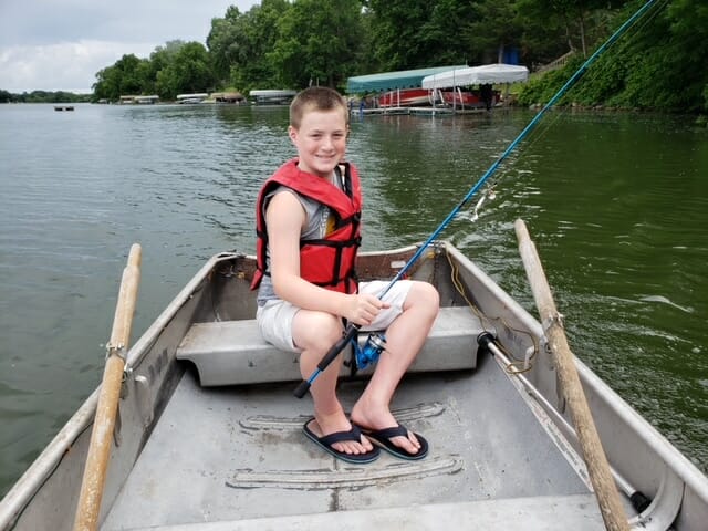 Caleb, in boat with fishing pole smiles brightly at the camera.