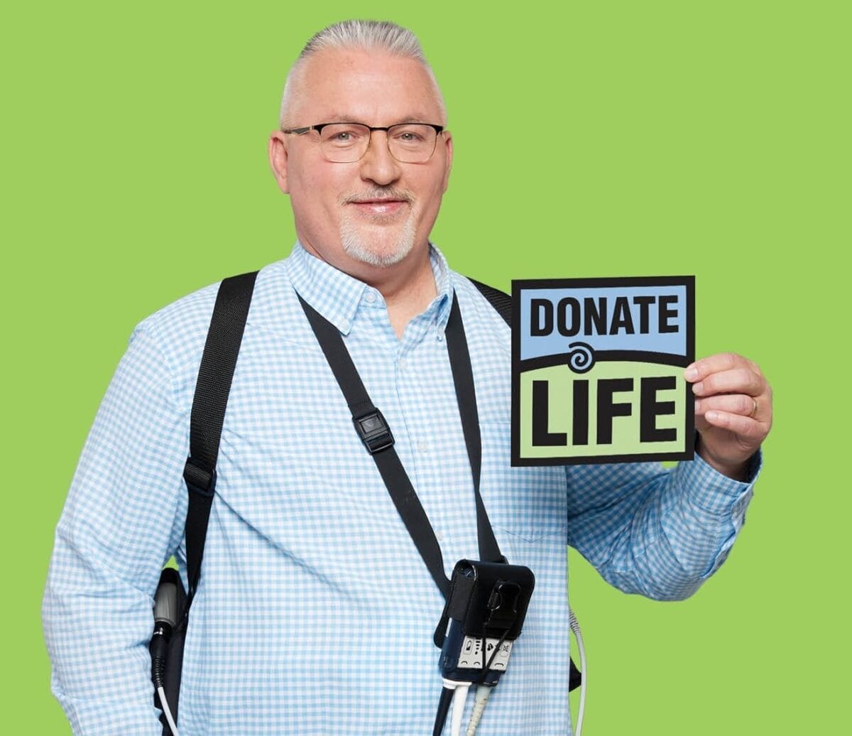 Man with glasses holds a DonateLife sign while wearing his LVAD machine.